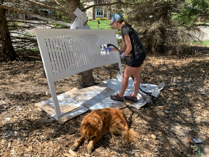 woman painting headboard with paint sprayer with dog on the ground